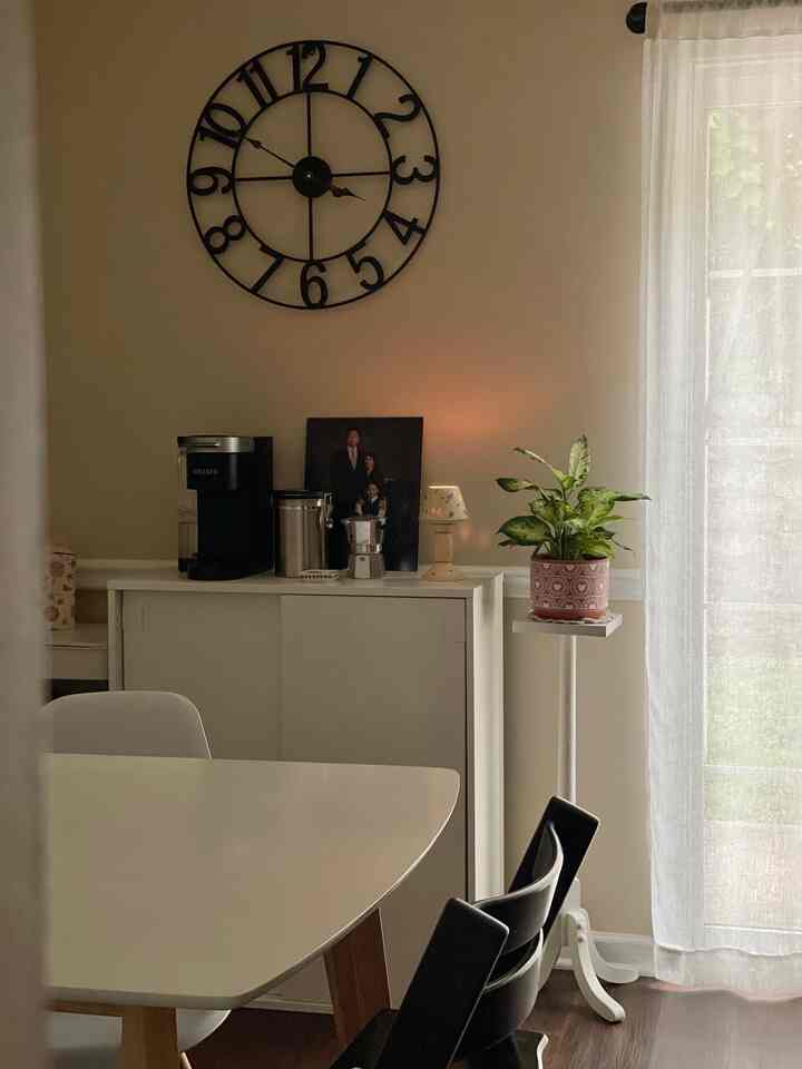 Beige and white toned dining room featuring a white dining table, black and white chairs, wall clock, and a green plant creating a calm atmosphere