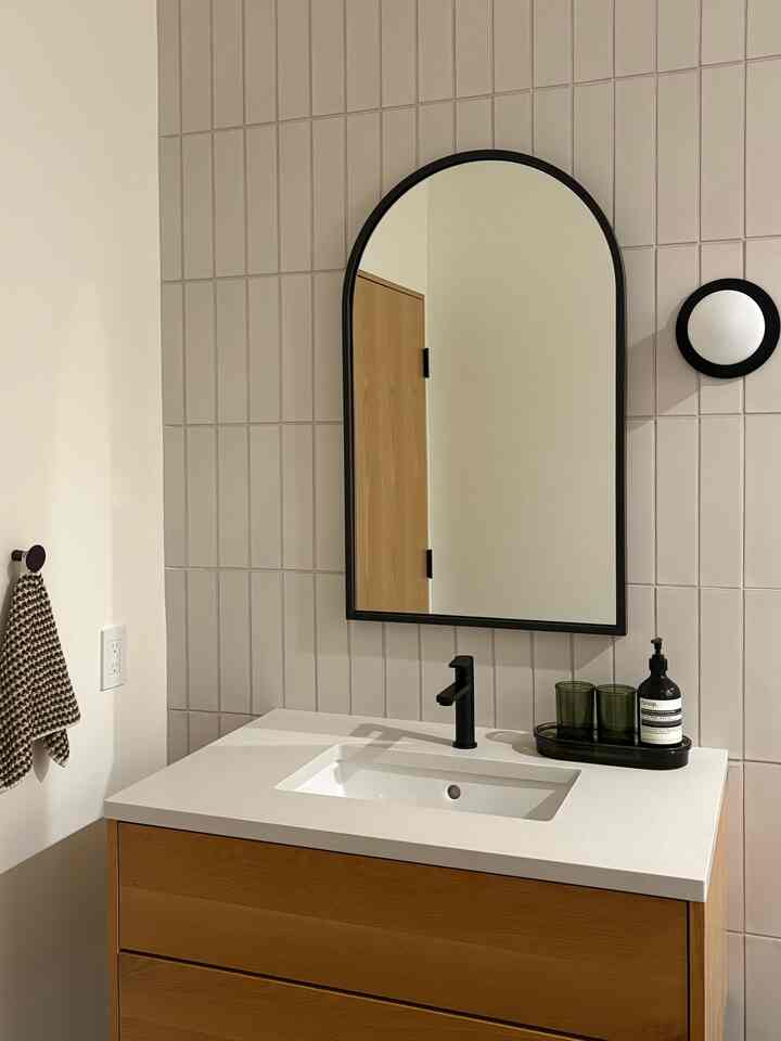 A modern and clean bathroom featuring white and wood tone vanity, black-framed arch mirror, and a simple integrated sink