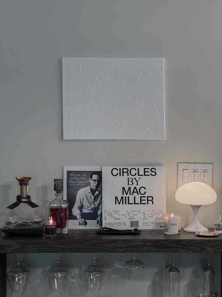 Minimal white and wood-toned home bar featuring candles, a modern table lamp, and vinyl record covers with cozy ambiance