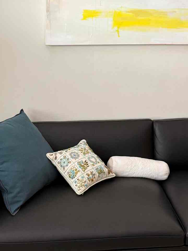 Modern living room space with a black leather sofa featuring assorted cushions, set against a white wall background with a bold yellow abstract painting