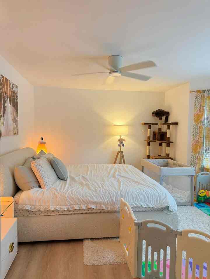 Bright white and natural toned bedroom-living room featuring a large bed, baby crib, cat tower, and baby playpen creating a cozy atmosphere