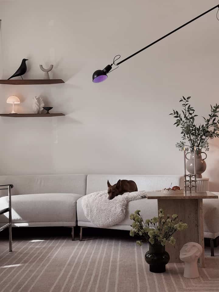 White and beige toned living room featuring a minimalist sofa, side table with flowers, wall lighting, and cozy decorative elements