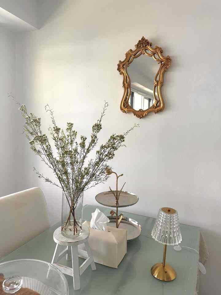 White and gold toned dining room featuring a plant stand on the table and an ornate mirror, presenting a simple aesthetic