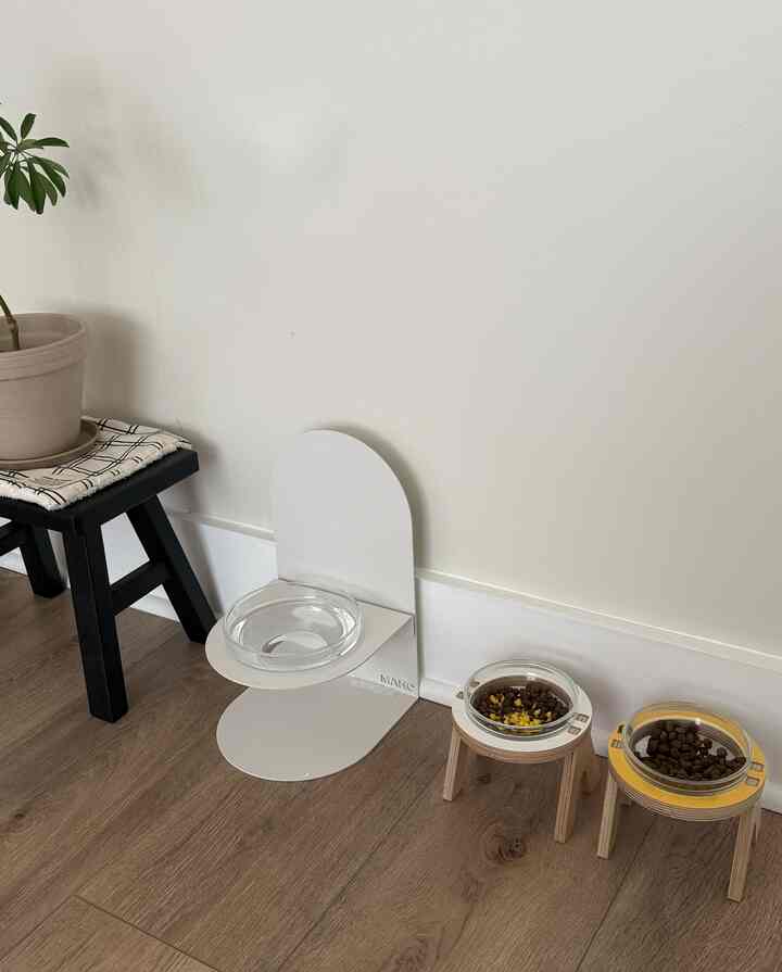 A clean pet feeding station in white and wood tones, featuring a small wooden stool and three pet food bowls arranged neatly.