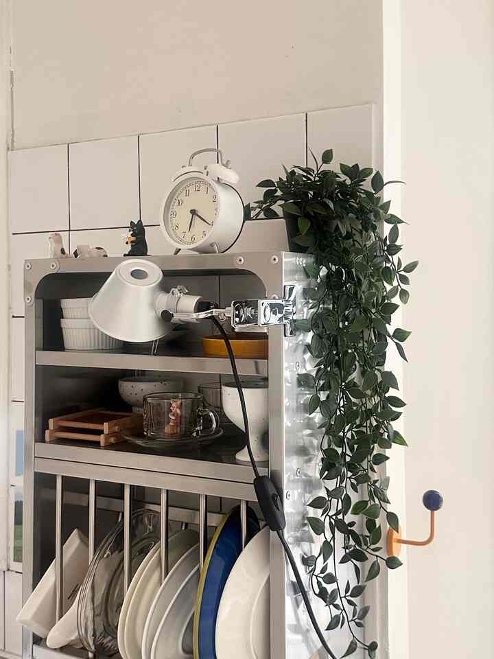 A cozy kitchen space with white and silver tones featuring a metal shelf with various plates, a clock, and cascading green plant