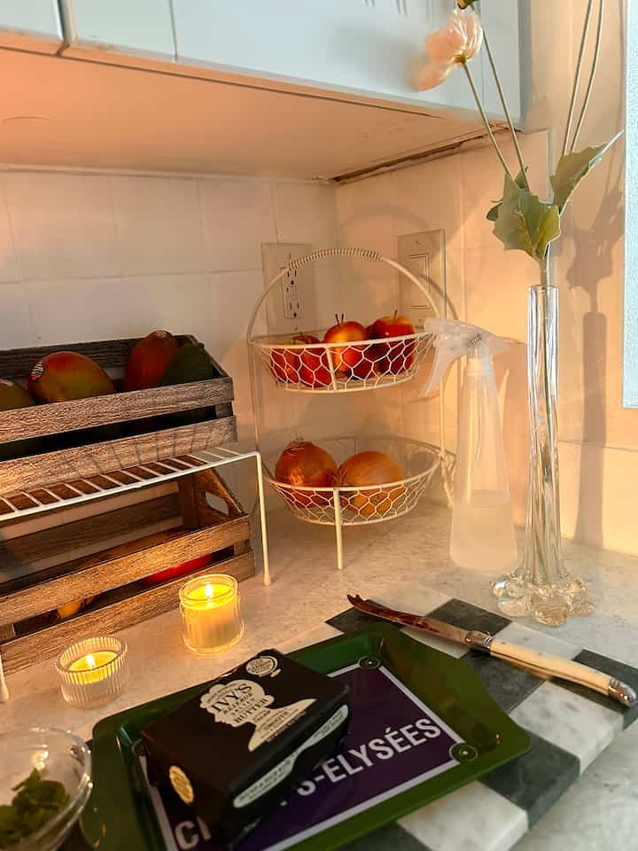 Cozy kitchen corner with white tiled wall, wooden crates holding fruits, tray with knife, fruit baskets, and candles on countertop