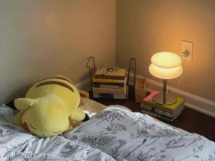 Beige-toned small bedroom featuring floral duvet cover, Pikachu plush, and glowing table lamp creating a cozy atmosphere