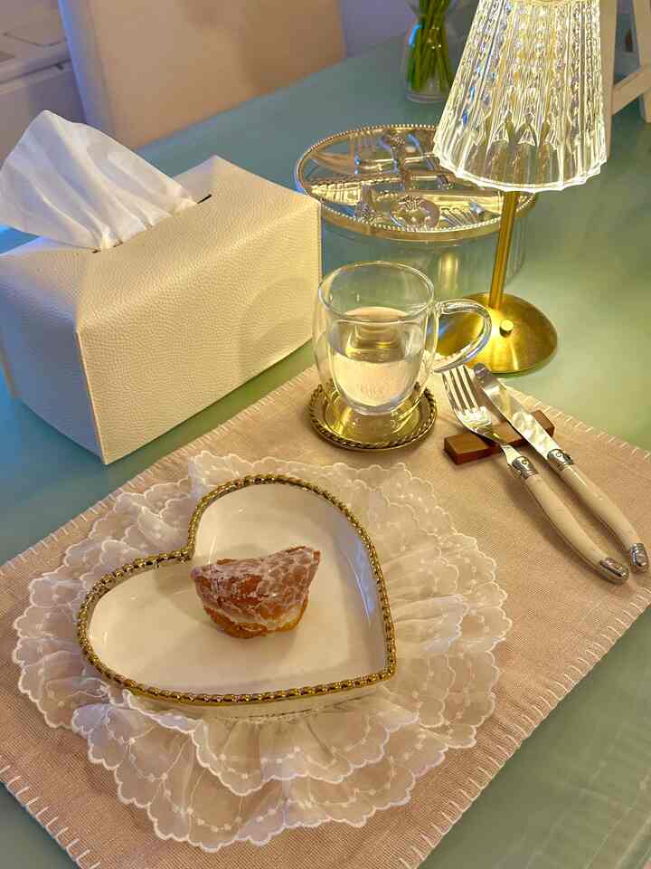 Beige and gold toned table setting featuring a lace-embellished tablecloth, heart-shaped dish with pastry, gold cutlery and clear glass cup, creating a cozy dining vignette