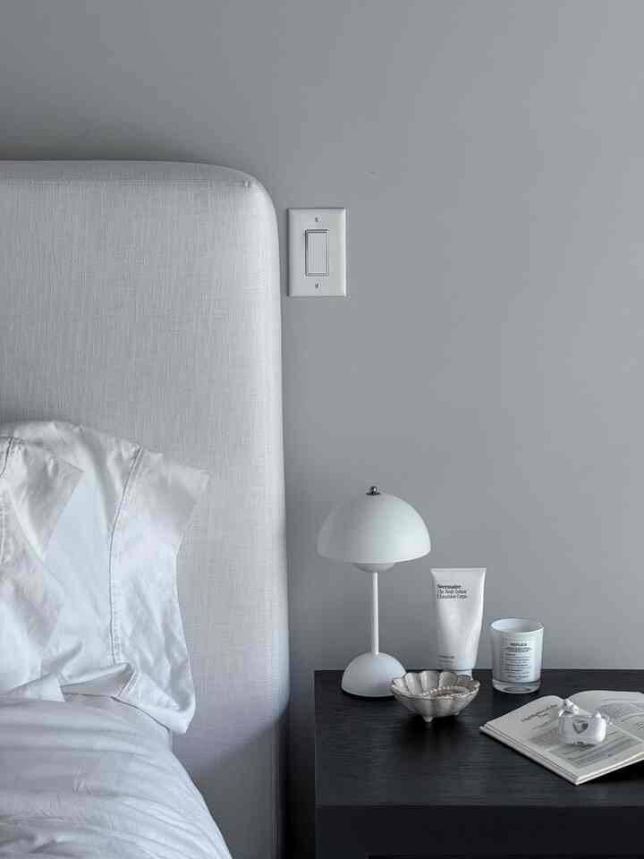 White and gray toned bedroom featuring a white bed, nightstand, table lamp, and candle, creating a clean minimal space