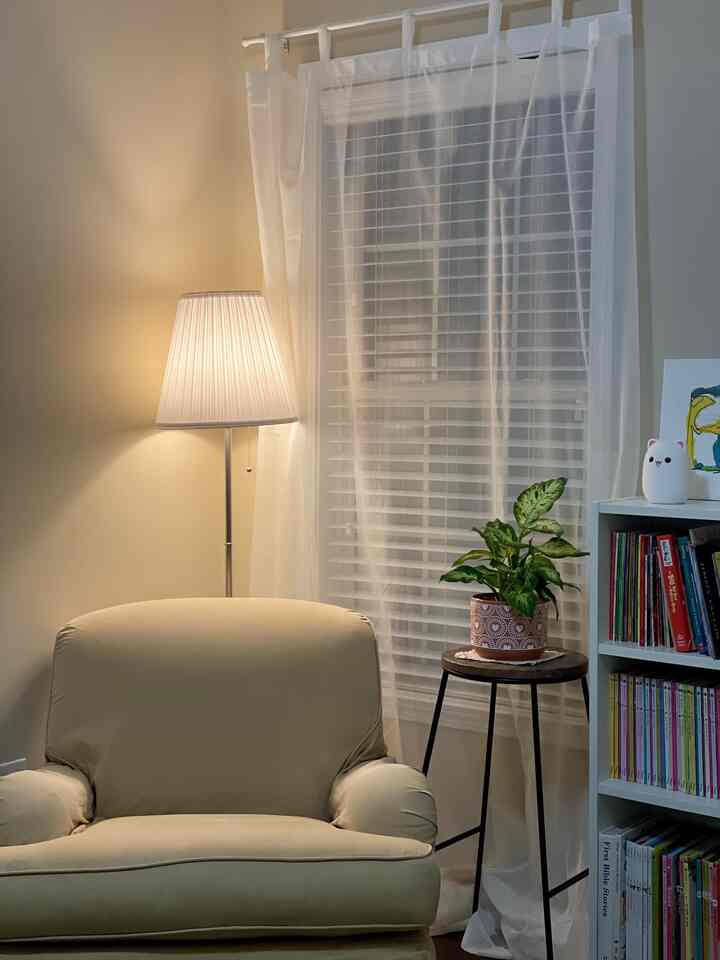 A cozy living room corner in white and beige tones featuring a beige armchair, floor lamp, small side table with plant, and a bookshelf filled with books
