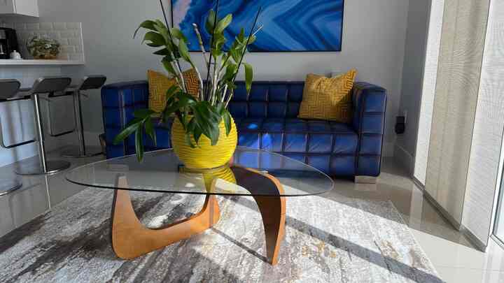 This tufted blue couch brings a pop of color to a postmodern-inspired interior, featuring a unique glass and wood coffee table and a bright yellow vase #cozyliving #livingroomstyle #homedecor #PostmodernDesign #BlueCouch