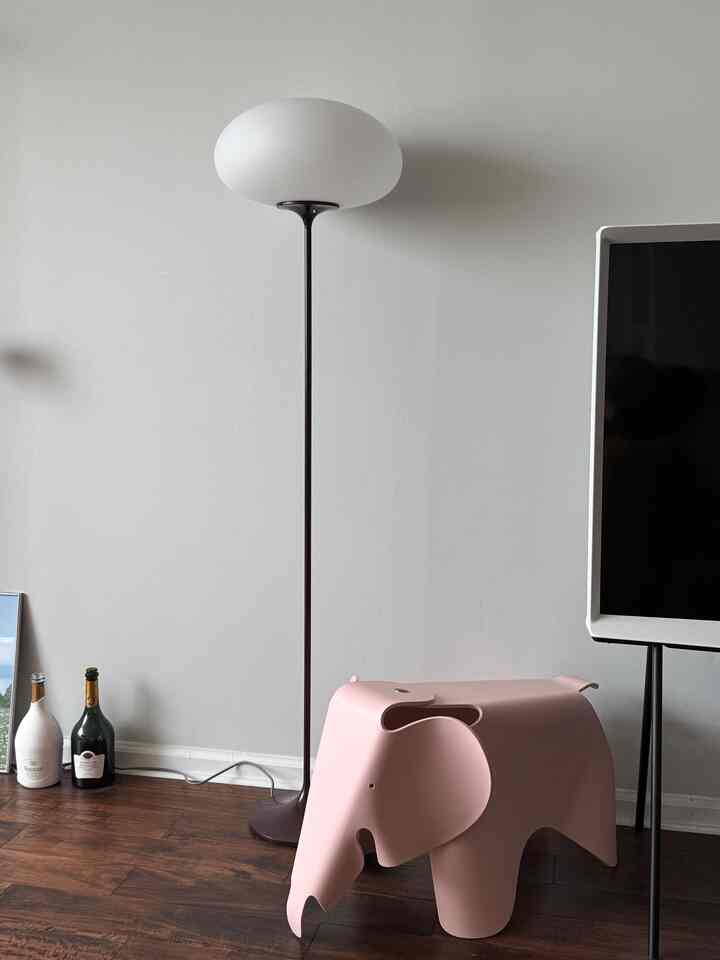 A modern living room with white walls and brown hardwood floors featuring a pink plastic elephant chair and a floor lamp
