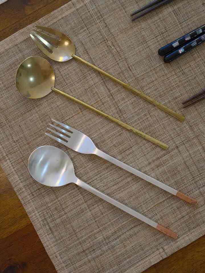 A simple dining setup photo showing gold and silver tone cutlery (spoons and forks) arranged on a brown wooden table with a woven placemat