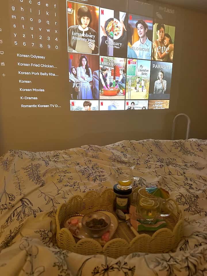 A cozy bedroom in white and natural tones featuring comfortable bedding and a wall projector for home cinema