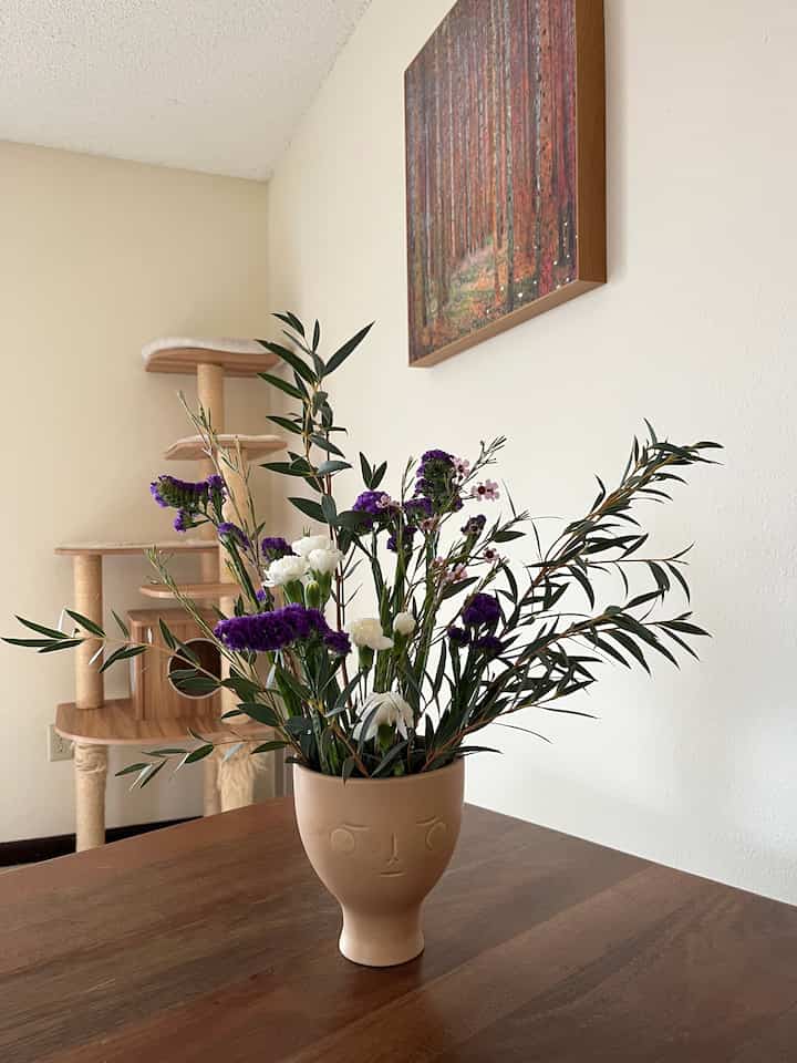 Beige-walled living room with wooden dining table featuring a vase of flowers and a cat tower, creating a natural ambiance