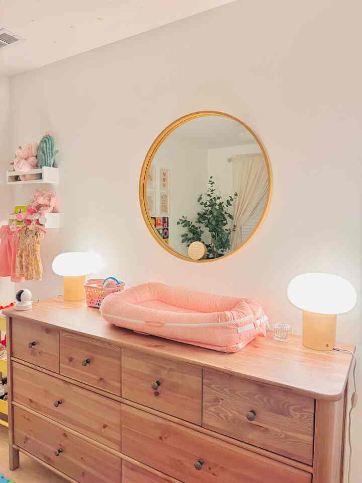 Natural-toned kids' room featuring a large round mirror and wooden dresser topped with pink baby items and soft table lamps creating cozy atmosphere