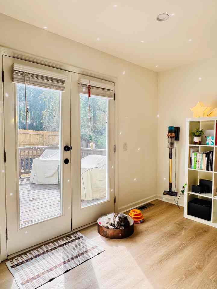 Bright white-walled living room with wood-tone flooring, featuring a cat resting in a brown scratcher, bookshelf and cordless vacuum present in cozy space