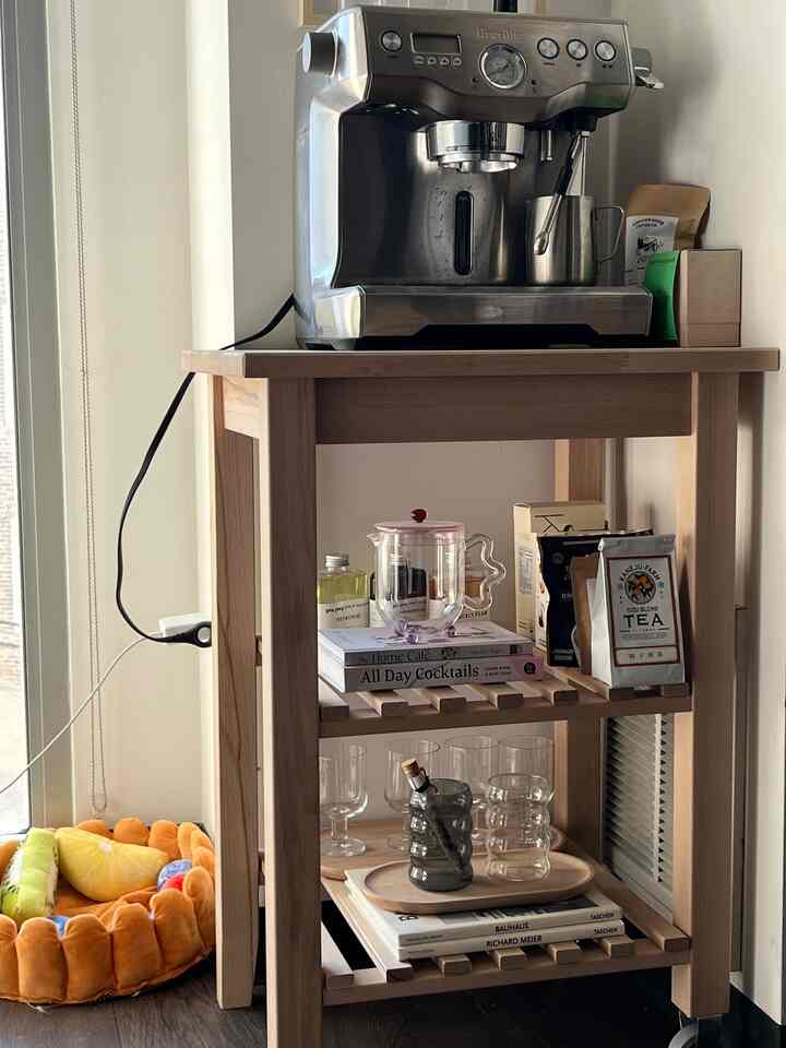Natural wood-tone kitchen cart with a silver coffee machine and transparent glasses in a home cafe setting, with an orange pet bed at bottom right