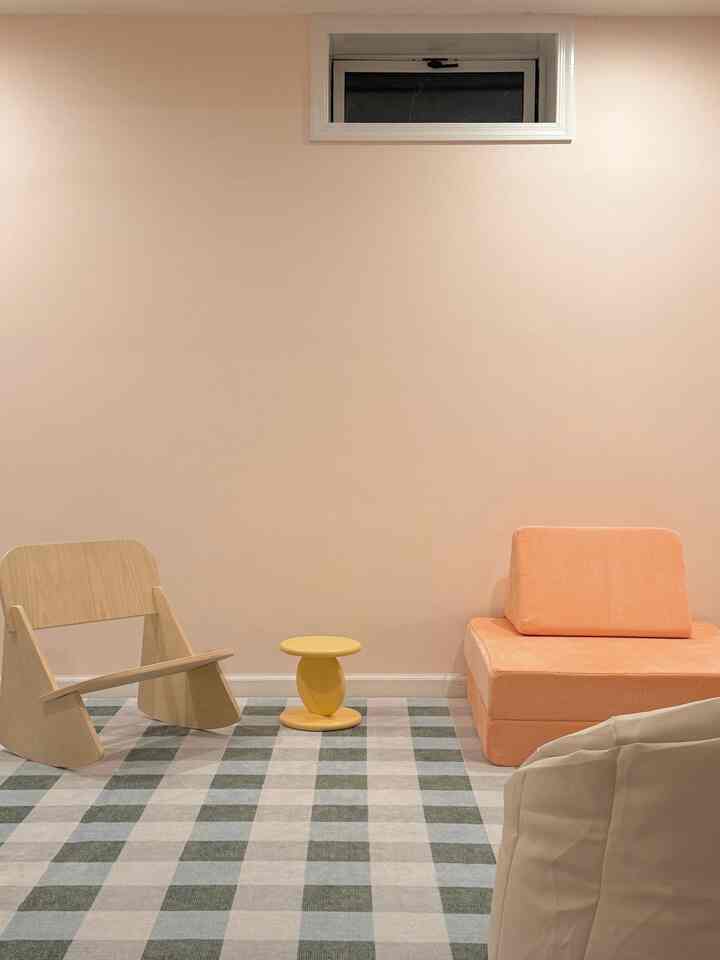 A kids' room with soft beige walls and checkered rug, featuring a minimal wooden rocker, a yellow stool, and an orange child-sized sofa in a cozy setting