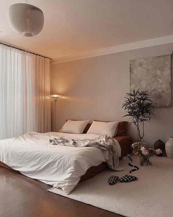 Cozy beige and white toned bedroom featuring a comfortable bed, floor lamp, and plant in a natural modern style space