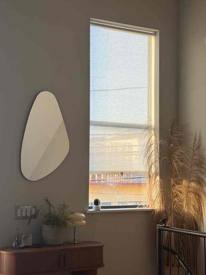 Cozy space with warm wood tone furniture, beige blinds on balcony window, a uniquely shaped mirror, and indoor plants