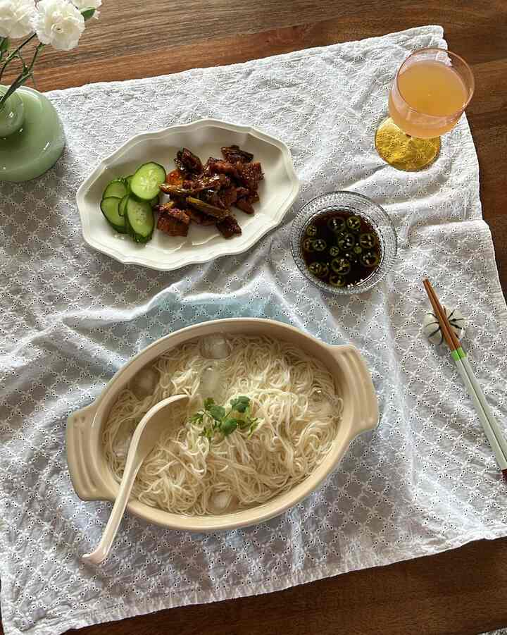 A home cafe table with beige ceramic bowl of noodles and side dish on a white lace table runner, accompanied by a yellow-stemmed wine glass