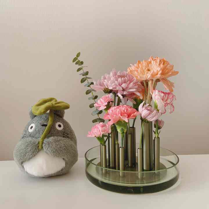 Natural color toned indoor space featuring a Totoro plush doll beside a glass vase with multiple pink and orange flowers, creating a cozy atmosphere
