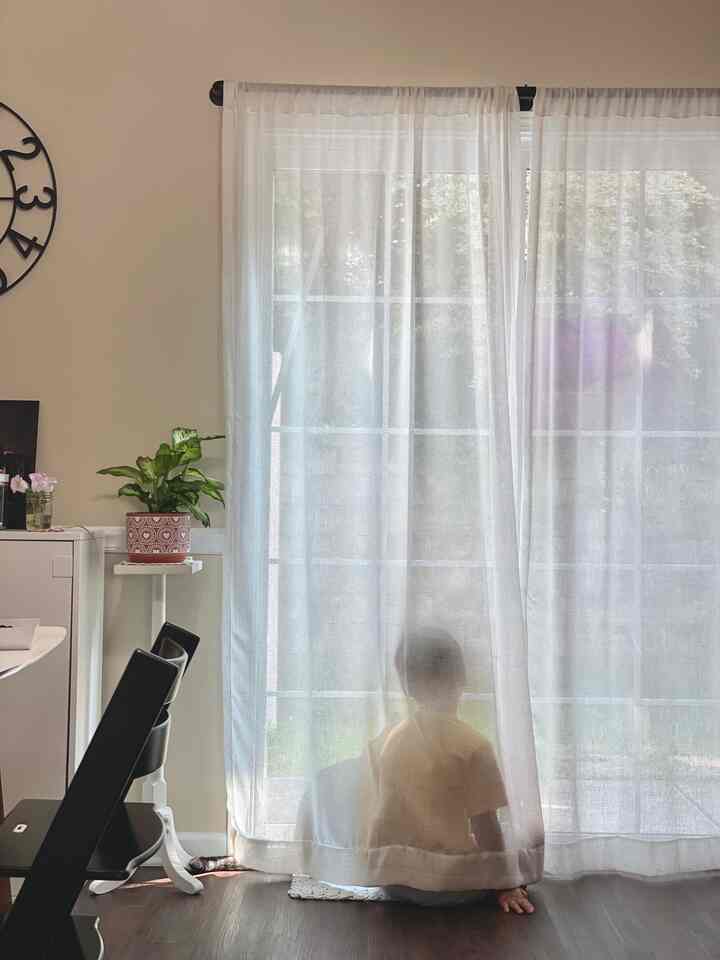 Bright white-toned living room with sheer white curtains and dining chairs, cozy space with a child and a dog by the window