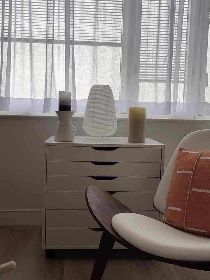 Minimal white-toned corner featuring a chest of drawers with paper table lamp and soft cushion giving a calm, clean atmosphere