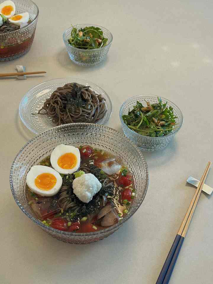 White table surface featuring transparent glass bowls and plates arranged with cold soba noodles and salad, creating a cozy summer dining setup