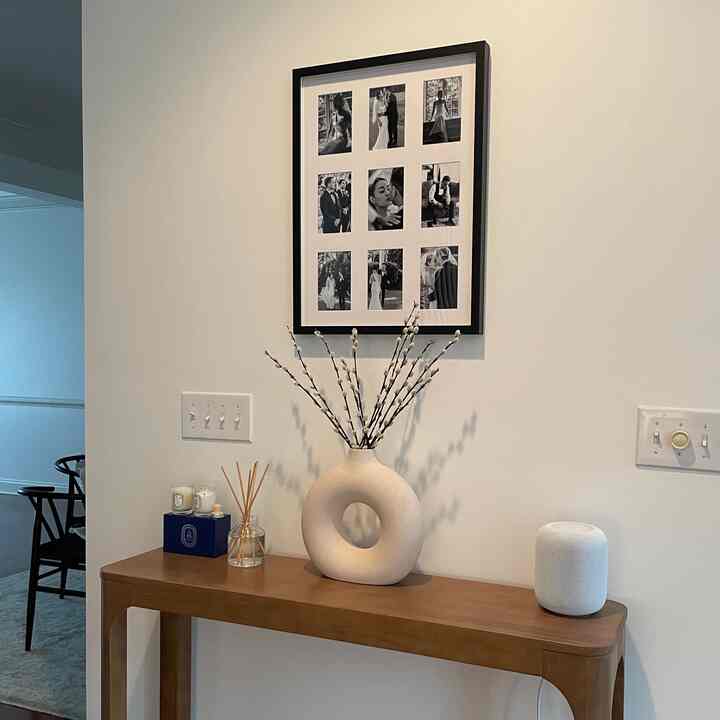 Beige-toned wall featuring a black framed photo collage, wooden console table topped with ceramic vase, diffuser, candles, and white smart speaker in a minimal natural living space