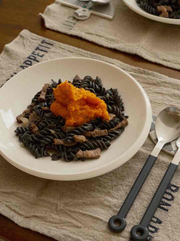 Beige tablecloth on wooden dining table with white plate featuring black pasta topped with orange kabocha cream in a simple setting