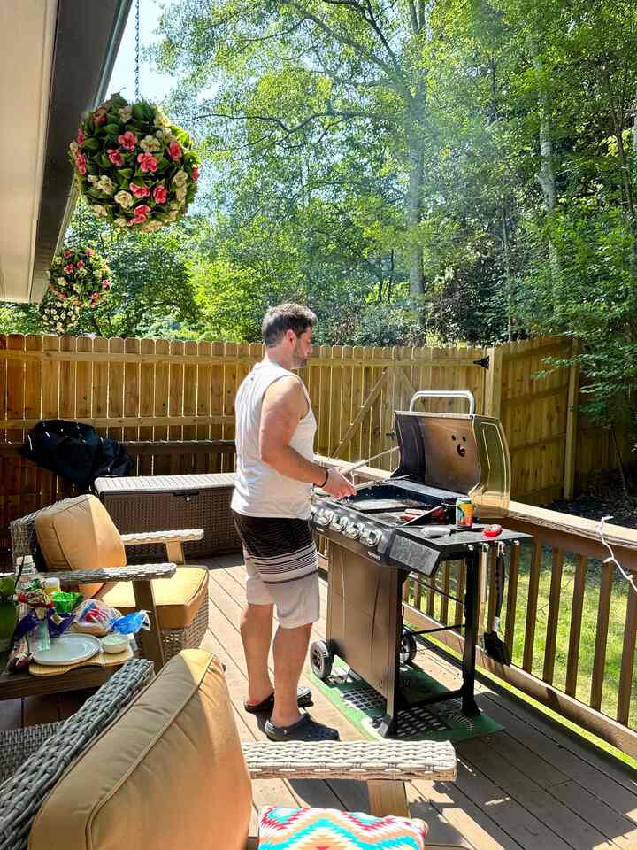 Outdoor balcony featuring green plants and brown wooden deck, beige cushioned outdoor chairs and grill create a cozy home party space