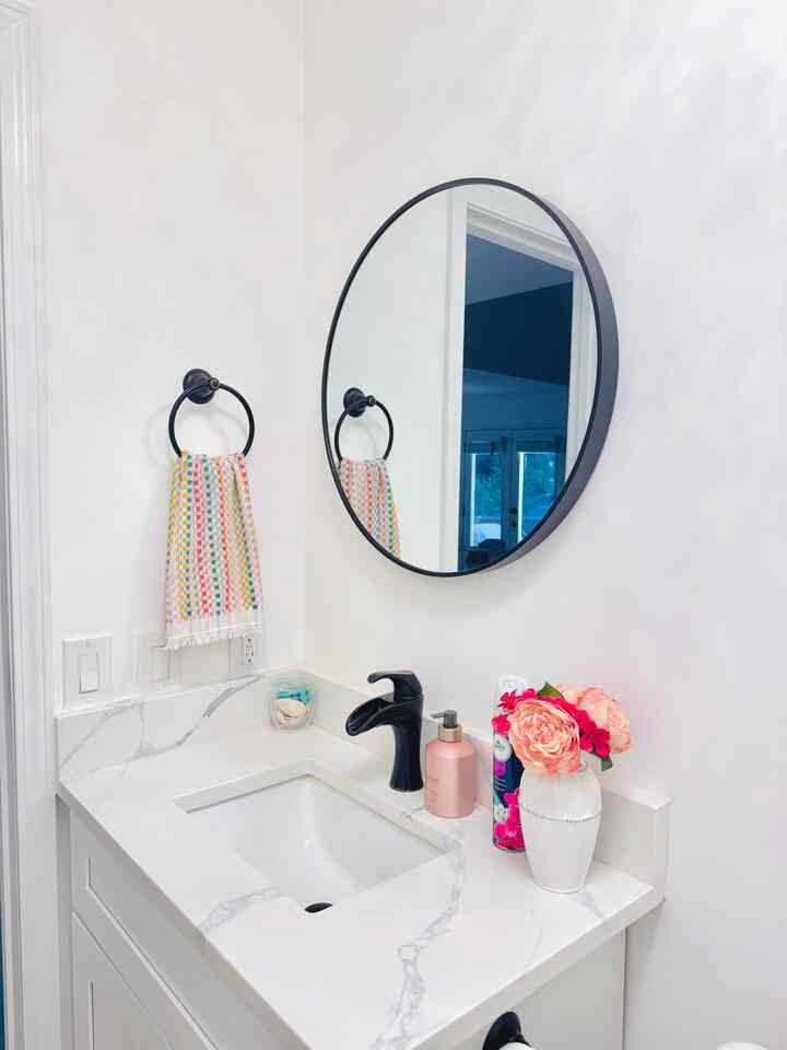 White-toned bathroom sink area with round mirror, pink vase flowers, and black faucet creating a simple, clean atmosphere