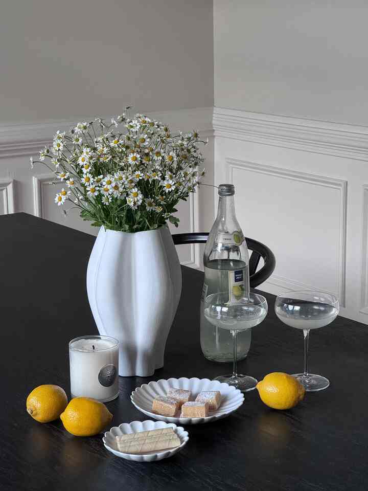 Black dining table in dining room featuring a white vase with fresh daisies, bottle with label, and two clear coupe glasses presenting a clean, elegant setup