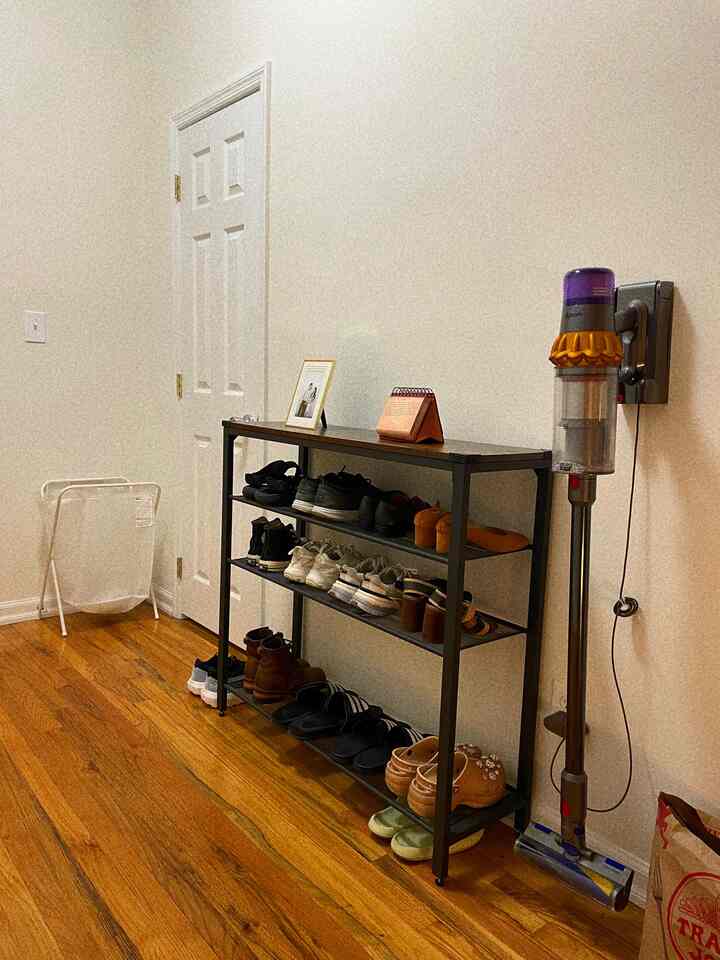 White-walled entrance with brown wooden floor featuring black metal shoe rack, cordless vacuum, and laundry hamper in neat arrangement