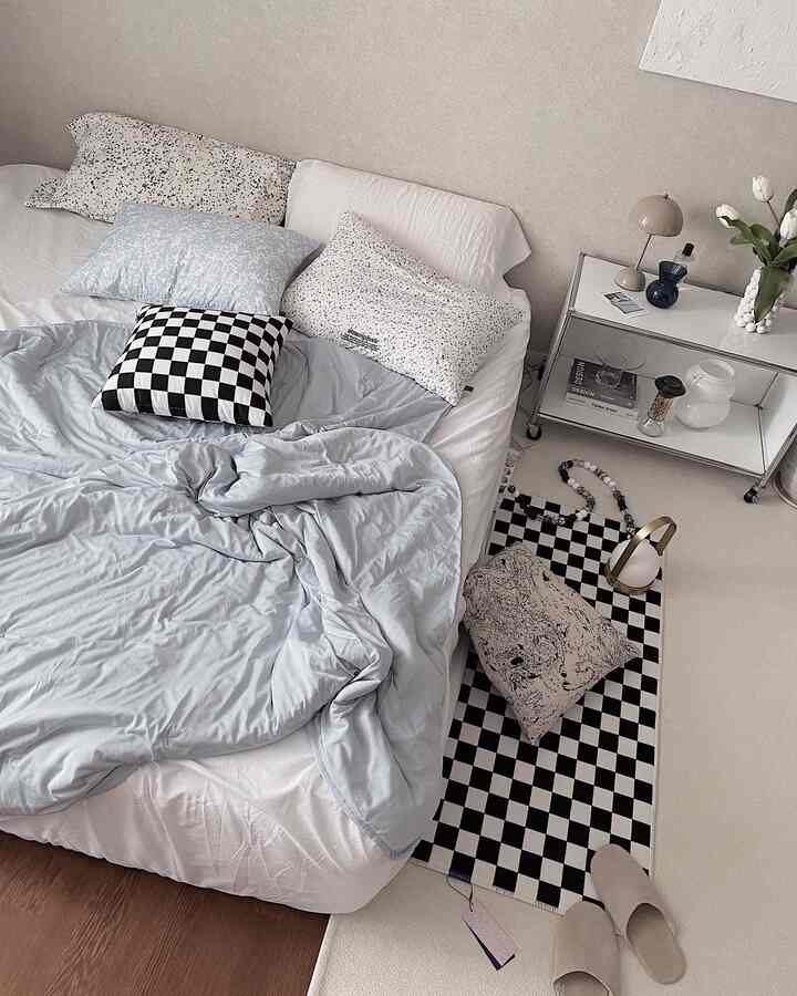 A white and sky blue toned bedroom featuring a cozy bed, checkerboard rug, side table, and minimalist relaxed atmosphere