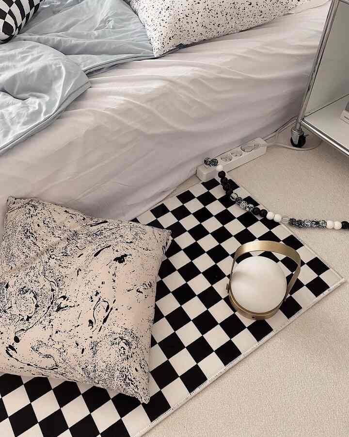A bedroom in white and black tones, featuring a checkered rug, patterned cushion, and portable table lamp creating a cozy space