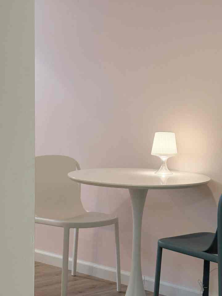 Small dining space in white and pink tones featuring a round table, table lamp, and two chairs with a simple atmosphere