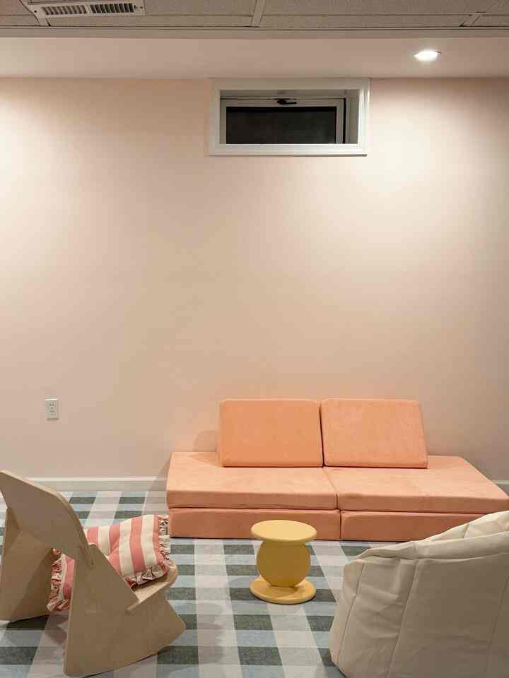 A cozy small room in beige and peach tones with a modular sofa, wooden armchair, and yellow stool positioned beneath a small high window