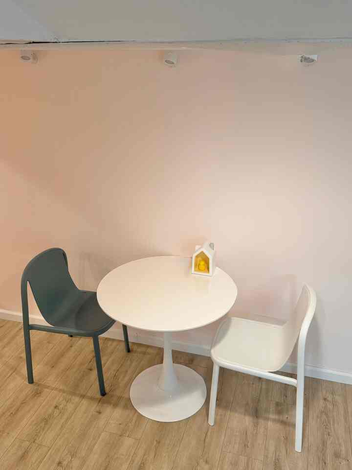 Soft pink wall and natural wood flooring complement a two-person white round dining table with simple chairs, creating a minimal and natural ambiance