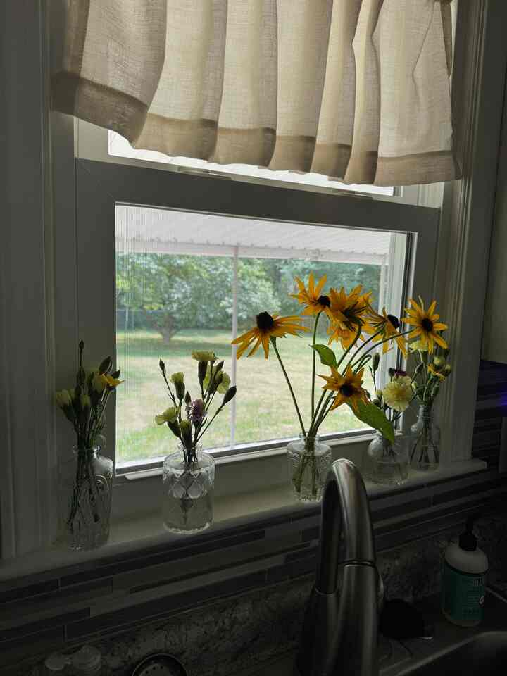 Kitchen window with soft beige linen curtains featuring transparent glass vases holding yellow sunflower flowers