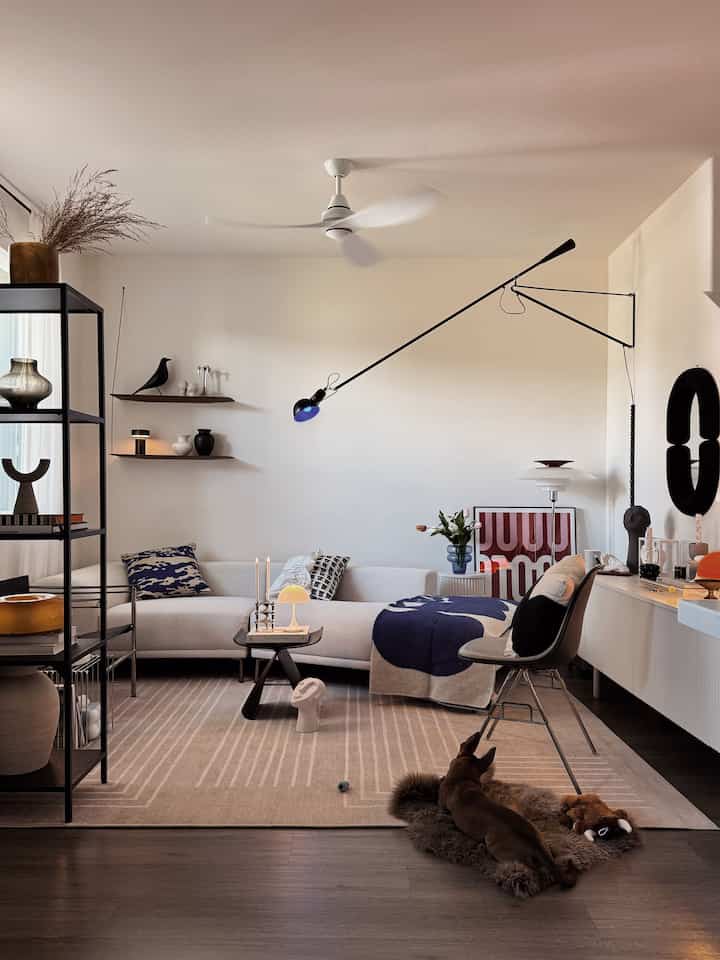 白とベージュのトーンのリビングルーム、中央に白いセクショナルソファとベージュのラグが配置され、前方に犬が寝そべり、ブルーのアクセントのスローや洗練されたミッドセンチュリーモダンの照明が調和した雰囲気のあるインテリア