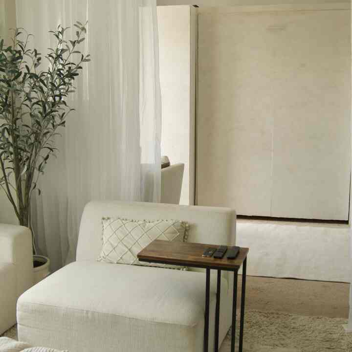 White and beige toned small living room featuring a simple single armchair, wooden side table, and sheer curtains creating a cozy atmosphere