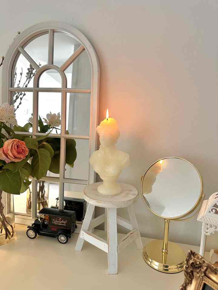 White-toned interior space featuring an arched window-style wall mirror and a lit bust-shaped candle on a distressed stool, creating cozy decor ambiance