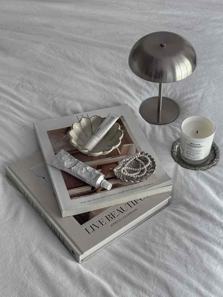 A monotone and silver-themed tabletop featuring books, trays, a scented candle, and a table lamp with a calm atmosphere