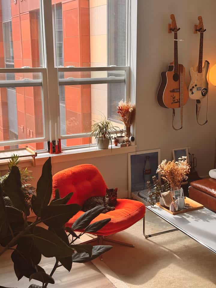 Bright orange armchair with orange exterior beyond window, natural-style living room interior featuring plants and guitars