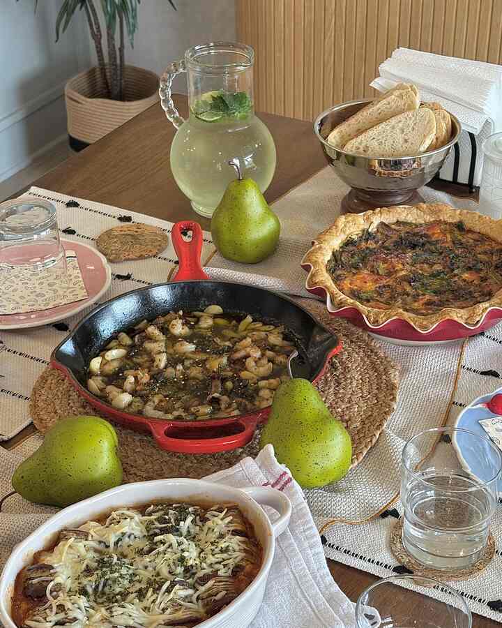 Natural wood-toned kitchen dining table featuring baked dishes, plates, glasses, and beige table runners creating a cozy atmosphere