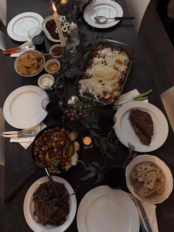 Black dining table set with white plates, diverse dishes, candles, and greenery for a cozy home party dinner setting