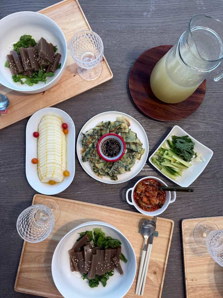Natural wood tone dining table featuring Korean side dishes, pajeon pancake, and drinks in cozy kitchen lunch setting